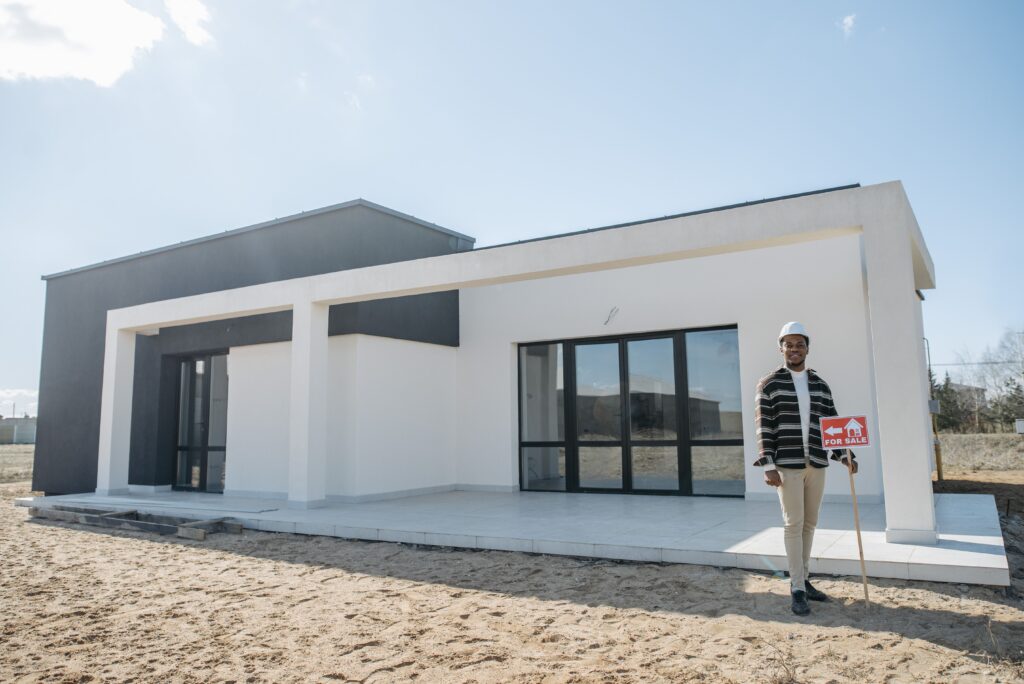 Man wearing a hard hat and a plaid jacket stands next to a modern white and gray house with a red 'For Sale' sign in front of it on a sunny day.
