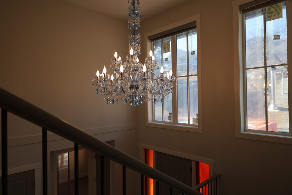 Crystal chandelier hanging above a stairwell, lit bulbs casting warm light by large windows in a neutral-colored interior.